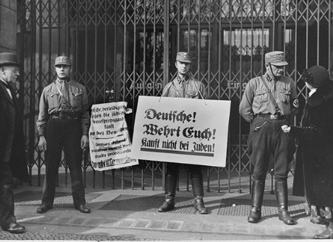 Three uniformed Storm Troopers with light skin tone stand in front of a closed shop. The soldier in the center holds a boycott sign in German, while another sign hangs from the gates covering the shop entrance. The soldier to the right appears to be speaking to a feminine figure with light skin tone in dark clothes.