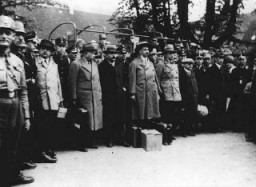 A group of men stand together. Most are wearing hats and outerwear. Uniformed guards stand around the perimeter of the group.